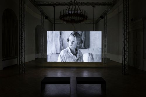 In einem Ausstellungsraum wird ein Film in projiziert. Er zeigt eine Frau mit halblangem blonden Haarschnitt in schwarzweiß