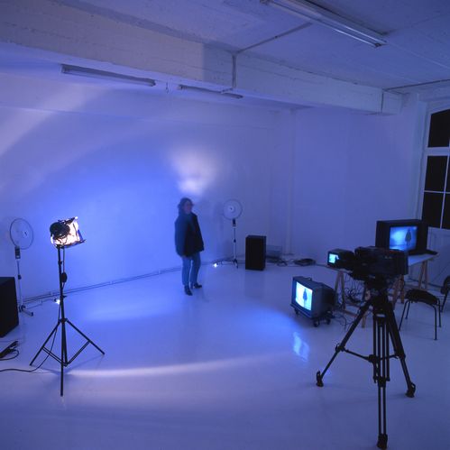A person stands in a darkened room in front of a camera and other technical devices such as spotlights and screens. The screens reproduce this image.