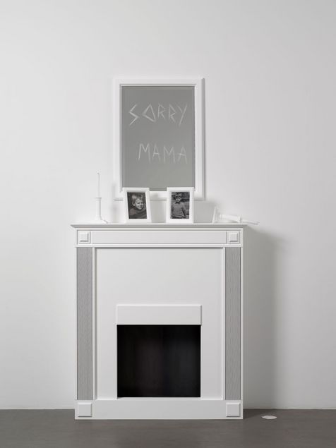 Above a neoclassical, entirely white fireplace hangs a white-framed mirror inscribed with “Sorry Mama” in angular lettering. On the mantelpiece sit two black-and-white childhood photographs and two white candlesticks, one of which has fallen over. The photographs depict Michael Elmgreen and Ingar Dragset.