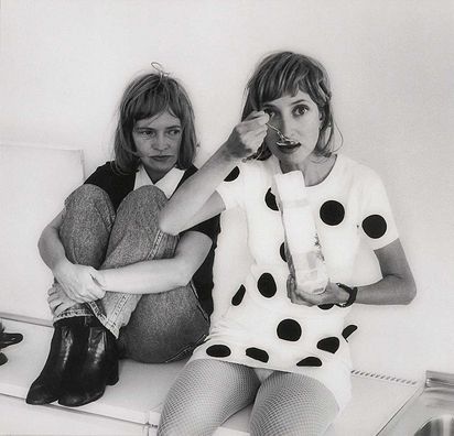 Eine Schwarz-Weiß-Fotografie zweier Frauenfiguren nebeneinandersitzend, die rechte isst etwas aus einer weißen Verpackung und sieht die Betrachtenden an, die linke Frau blickt sie an. Die Szenerie und ihre Proportionen haben haben irritierende Wirkung.