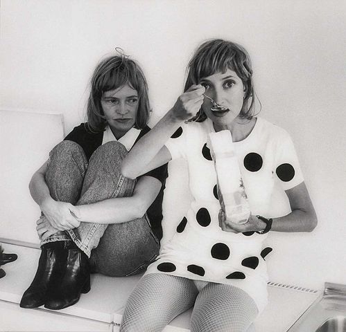 Eine Schwarz-Weiß-Fotografie zweier Frauenfiguren nebeneinandersitzend, die rechte isst etwas aus einer weißen Verpackung und sieht die Betrachtenden an, die linke Frau blickt sie an. Die Szenerie und ihre Proportionen haben haben irritierende Wirkung.