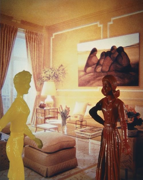Against a yellow-toned background, two slender doll figures are photographed. They are made of plastic, have a glossy surface, and are monochrome in yellow and brown. The photograph forming the backdrop shows an elegant yellow-toned living room with panelled walls and select modern furnishings.