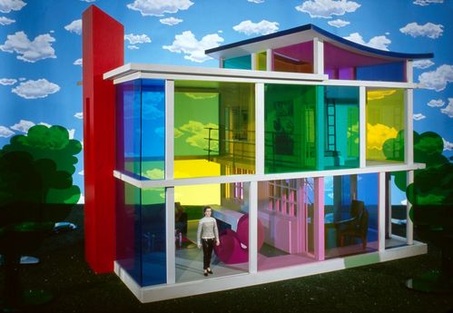A modernist dollhouse stands in front of a blue-and-white cloud-patterned wallpaper. Plexiglas shrubs frame the house. It has three floors, and the Plexiglas windows are in various highly saturated colours. At one of the large ground-level windows leading into the garden stands a woman in a striped T-shirt.