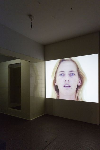 In einem weiß gekachelten Raum projiziert ein Projektor auf einen Bereich ungekachelter Wand. Zu sehen ist eine Nahaufnahme des Gesichts einer blassen Person mit langen blonden Haaren.