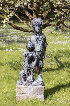 A bronze-colored sculpture stands on a rectangular stone base outdoors on a grassy lawn. The figure depicts a standing person in shorts and a loose top, resting one hand on a dog or animal-like figure. In the background, a tree with small white blossoms is in bloom.