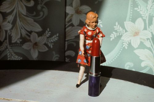 In front of a mint-colored floral wallpaper stands a doll figure before an oversized lipstick. The red floral dress of the doll figure and the red of the lipstick harmonise with each other.