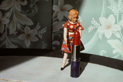 Vor einer minzfarbenen, geblümten Tapete steht eine Puppenfigur vor einem überdimensionalen Lippenstift. Das rot geblümte Kleid der Puppenfigur und das Rot des Lippenstifts harmonieren miteinander.
