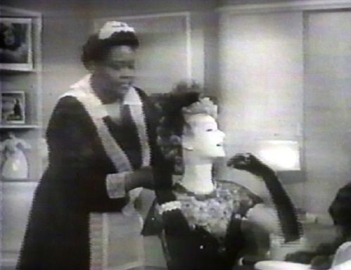 Stereotypical film scene of an elegant white woman sitting at a dressing table while a plump black maid does her hair.