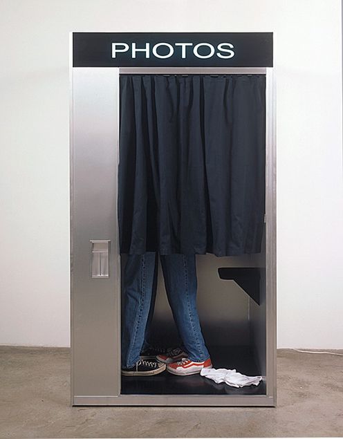 A silver aluminium photo booth stands in front of a white wall. Behind the closed black curtain, only the legs of two people in jeans are visible, standing so close together that it gives the impression the pair are kissing.