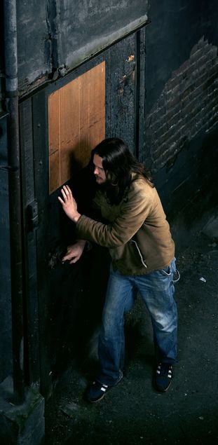 Zu sehen ist ein Mann mit schulterlangen Haaren, der eine Tür öffnet. Er trägt eine helle Jeans und eine braune Lederjacke. Boden, Wände und Tür sind schwarz, wodurch die Szene insgesamt sehr dunkel wirkt. In der Tür befindet sich ein rechteckiges Element aus hellem Holz, das sich deutlich vom dunklen Hintergrund abhebt. Am linken Bildrand ist ein Rohr zu sehen, das sich vom Boden bis zur Decke erstreckt.