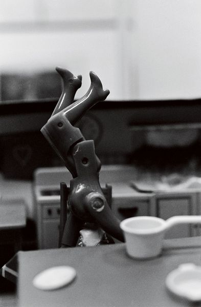 The black-and-white photograph shows a dollhouse kitchen in which a doll figure wearing a suit appears to be doing a handstand on a chair. In the foreground there is a set kitchen table, and in the background a kitchen unit. Both planes are heavily blurred, as the photograph focuses exclusively on the doll figure. The doll’s face is hidden beneath the table, and its legs are bent.