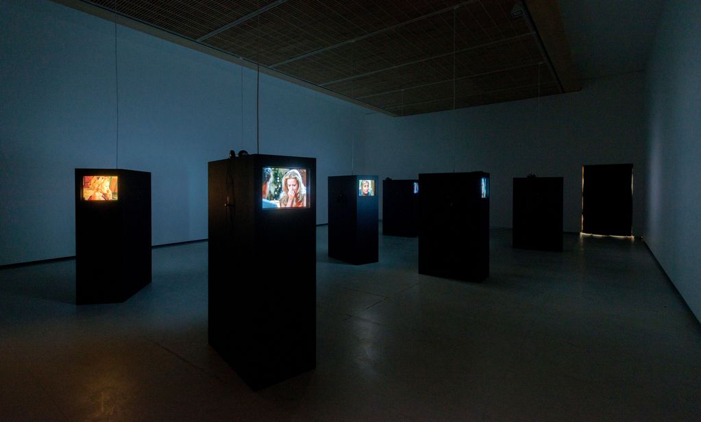 Ausstellungsraum mit 5 Monitoren auf schwarzen Sockeln. Auf den Bildschirmen laufen Spielfimszenen in Farbe.