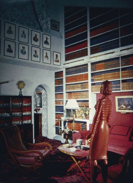 A slender, dark red lacquered doll figure stands facing a wallpaper backdrop. It depicts a library. Shelves are mounted on a very high wall, filled with uniformly bound leather volumes. Several pictures are hung on the walls. A globe stands on one of the shelves, and the seating is upholstered in red and arranged around a small coffee table. The doll’s sweater features a distinctive cable-knit pattern.
