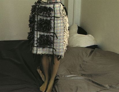 Der Bildausschnitt zeigt den mit Wollfäden applizierten Karorock einer jungen Frau, die auf einer grauen Lederlandschaft steht.