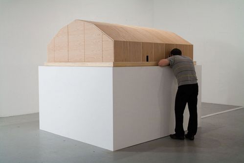 Architectural wooden construction on a plinth. A person looks through a small door into the interior of the sculpture