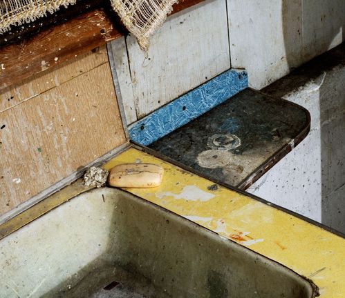 Zu sehen sind zwei unmittelbar nebeneinanderstehende Waschbecken. Beide weisen deutliche Gebrauchsspuren auf und sind mit Farbflecken verschmutzt. Das linke Waschbecken enthält ein Stück Seife sowie ein zerknülltes Papiertuch. Das rechte Waschbecken wirkt stärker abgenutzt und zeigt dunkle, schwarze Verfärbungen entlang der Ränder. Am oberen Bildrand ist ein Teil eines gehäkelten Tuches erkennbar.
