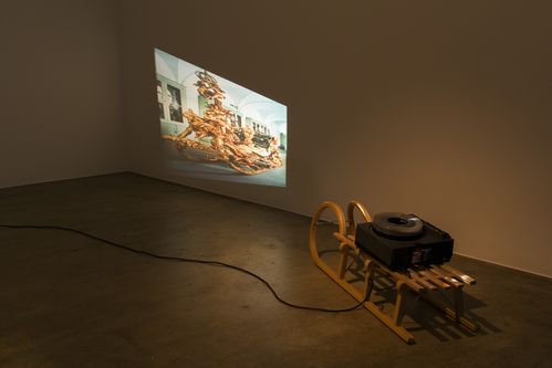 In one room, a slide projector stands on a wooden sled on the floor. It projects the image of a golden sledge onto the wall. The wooden sledge and the projector are directed obliquely against the wall, and the projection is distorted accordingly. 
