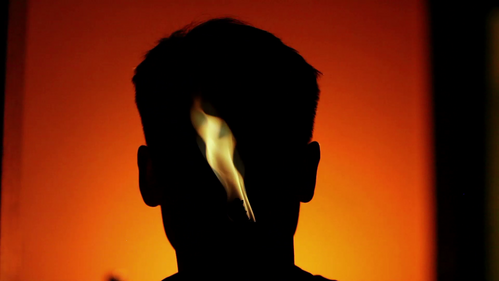 Video Still: Schattenbild eines menschlichen Kopfes mit kurzen Haaren vor hellrotem Hintergrund. In der Mitte des Kopfes brennt eine Flamme. 