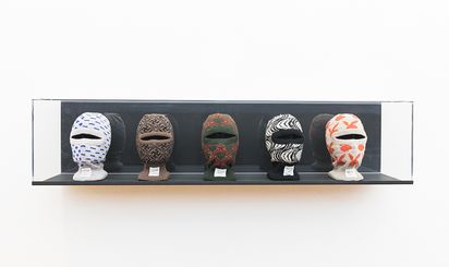 In einer länglichen, horizontalen Glasvitrine stehen fünf Puppenköpfe nebeneinander. Jeder trägt eine gestrickte Sturmhaube mit unterschiedlichem Muster. Von links nach rechts sind zu sehen: Plus- und Minuszeichen, Hakenkreuze, ein abstraktes, nicht eindeutig identifizierbares Muster, zebraartige Streifen sowie das Hasenlogo des Playboy-Magazins.