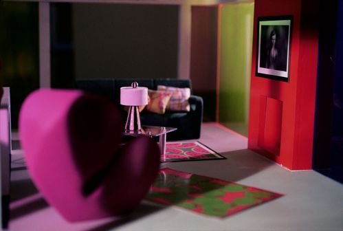 Shot frontally, the image shows a view into a modernist dollhouse. In the foreground stands a lilac wingback chair. Behind it are a coffee table placed on two colourful rugs and a black sofa. Above a red wall with an opening for a fireplace hangs a blurred black-and-white image of a female doll. The entire room is illuminated in red-purple light.