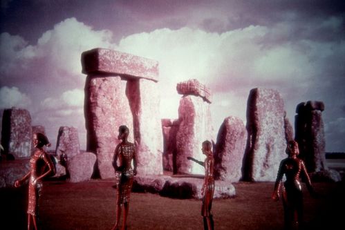 In front of a pink-toned Stonehenge backdrop, doll figures are standing. They are lacquered in brown and have a slender silhouette.