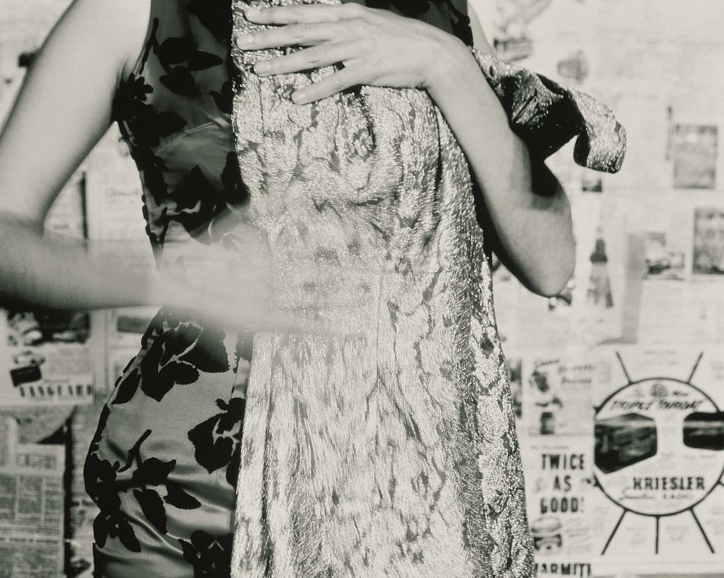 Black and white photograph of a woman in a dress with black roses printed on it, whose head is not visible, holding another dress to her body