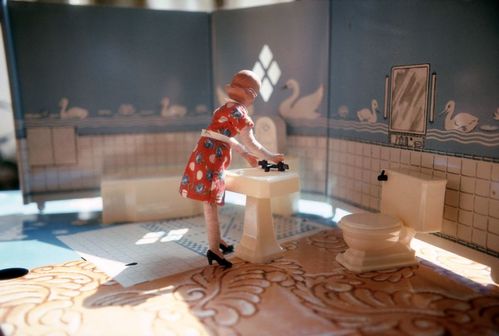 A doll figure wearing a red floral dress stands in the bathroom of a dollhouse. Tile patterns are printed on various sheets of paper. The figure stands beside a miniature sink. Bright light enters the room through a window from the side.