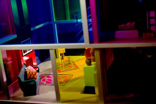 The coloured sliding windows of a  modernist dollhouse offer a view into its interior. Through the tinted glass, the living room is revealed, bathed in yellow light. The adjacent room is unlit and therefore barely visible.