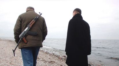 Two men walk side by side along the beach. One is carrying a machine gun on his back