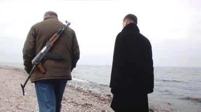 Zwei Männer gehen nebeneinander am Strand entlang. Einer trägt ein Maschinengewehr am Rücken