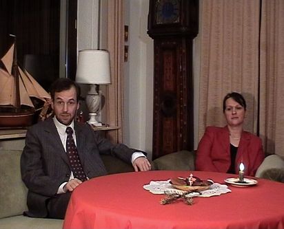 Zwei Menschen in festlicher Kleidung sitzen an einem Tisch mit roter Tischdecke. Auf dem Tisch stehen eine Kerze und ein einzelner Tannenzweig. Im Hintergrund sind schwere Vorhänge, ein Modellschiff und eine Lampe mit Marmorfuß zu erkennen.