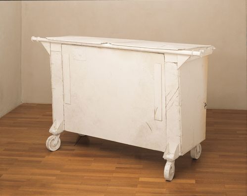 Eine einem US-amerikanischen Müllcontainer nachempfundene Skulptur steht auf einem Parkettboden. Sie ist wie ein echter Müllcontainer auf Rollen angebracht und vollständig weiß gehalten. Die Containerform ist mit verschmutzten Schaumplatten verkleidet, die Abdrücke aufweisen, als sei darauf gelaufen worden.