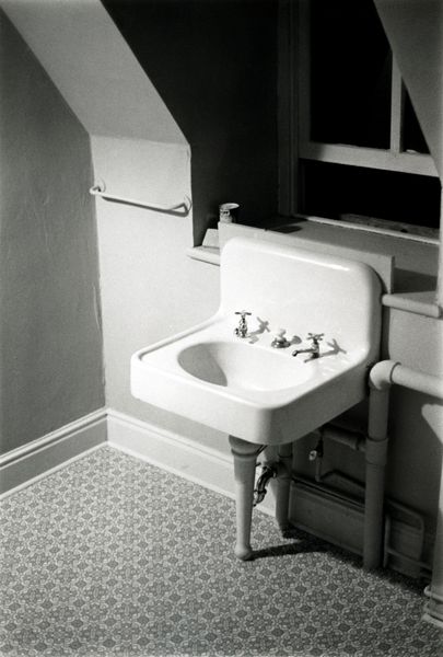 The black-and-white photograph shows a dollhouse bathroom. The frame focuses on a washbasin in front of a slightly open window. Next to the sink, realistic miniature pipes are installed.