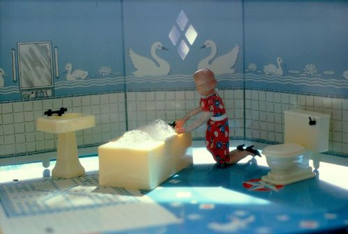A woman wearing a red floral dress is taking a bath. The dollhouse bathroom is tiled in white up to waist height and features blue wallpaper with a swan motif above.