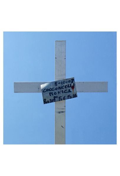 Photographs of white grave crosses against blue cloudless sky. All have simple memorial plates made of sheet metal, on which the names and life dates of the deceased are handwritten. Children's toys or stuffed animals are attached to many of the crosses. Both the crosses and the toys have been soiled by the weather and have come down. In some photos, wild grasses and yellow flowers protrude into the picture from below.