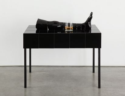 Auf einem schwarzen hochglänzenden Tisch liegt eine schwarze Keramikskulptur, die ein Bein darstellt. Daneben steht ein Glas mit einer Spirituose.