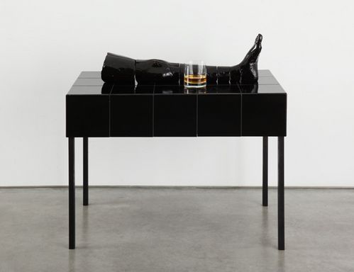 Auf einem schwarzen hochglänzenden Tisch liegt eine schwarze Keramikskulptur, die ein Bein darstellt. Daneben steht ein Glas mit einer Spirituose.