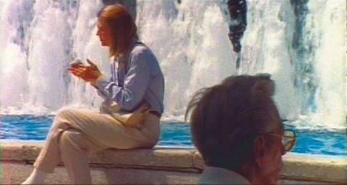 A woman wearing a blue shirt and beige trousers sits in front of a fountain. In the foreground of the image, the head of an elderly man wearing yellow-tinted sunglasses is visible.