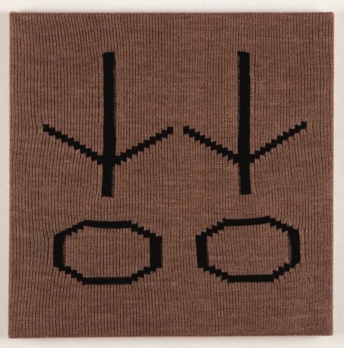 Ein auf Keilrahmen gespanntes gestricktes braunes Stoffstück mit zwei in schwarzem Wollstoff identischen Zeichen eingestrickt.