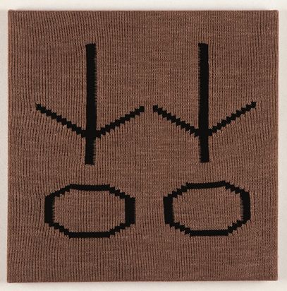 Ein auf Keilrahmen gespanntes gestricktes braunes Stoffstück mit zwei in schwarzem Wollstoff identischen Zeichen eingestrickt.