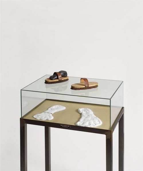 In einem Glasschaukasten mit hohen Tischbeinen liegen zwei gipsartige weiße Fußabdrücke. Auf der Glasabdeckung liegen zwei Handbürsten, in der linken steckt eine schwarze Gipsplastik eines Fußes.