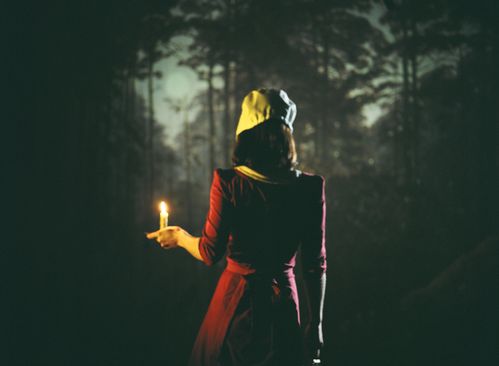 A young woman in a red dress and white hood holds a candle in her hand against the backdrop of a forest