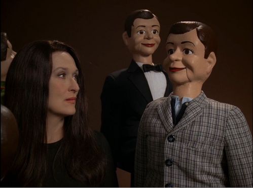 Die Schauspielerin Meryl  Streep mit schwarzer Perücke blickt seitlich auf eine Bauchrednerpuppe im karierten Anzug. Im Hintergrund ist eine weitere Bauchrednerpuppe im Smoking zu sehen.