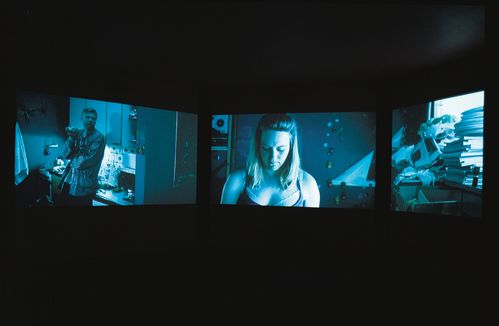 Three video projections side by side. On the left is a man in a kitchen, in the middle a woman, on the right a pile of books