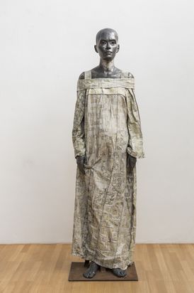 A three-dimensional sculpture of a woman in a long robe. The sculpture stands firmly on both feet, her arms hanging loosely at her sides. She looks directly at the viewer.