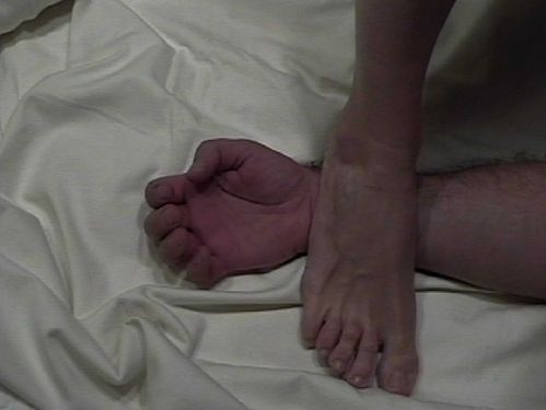 On a white bed sheet, a foot steps onto the wrist of another person. 