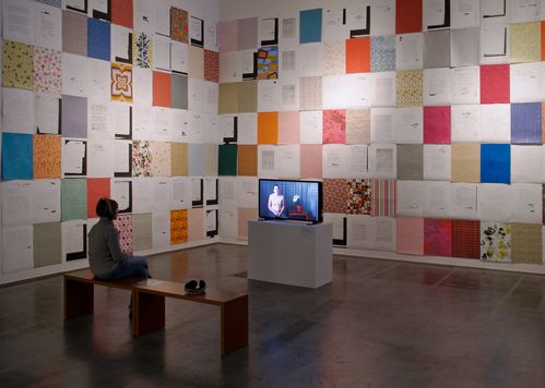 Installation with colorful paper rectangles across a corner on a wall. In front of it, a visitor with headphones is watching a video on a flat screen