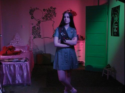 Eine junge Frau steht in einem Schlafzimmer, das pink und grün beleuchtet ist. Sie trägt eine Schärpe