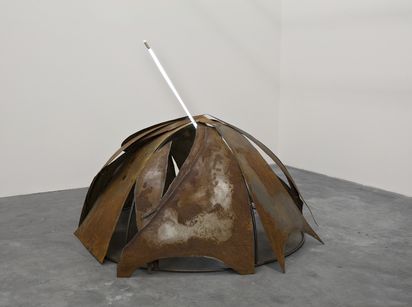 A dome-shaped rusted metal sculpture made of overlapping curved steel panels arranged like petals on a concrete gallery floor, pierced near the top by a glowing vertical white neon tube.