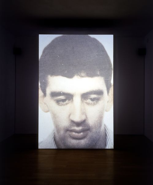 The face of a man with short dark hair and a lowered gaze is projected onto a partition wall